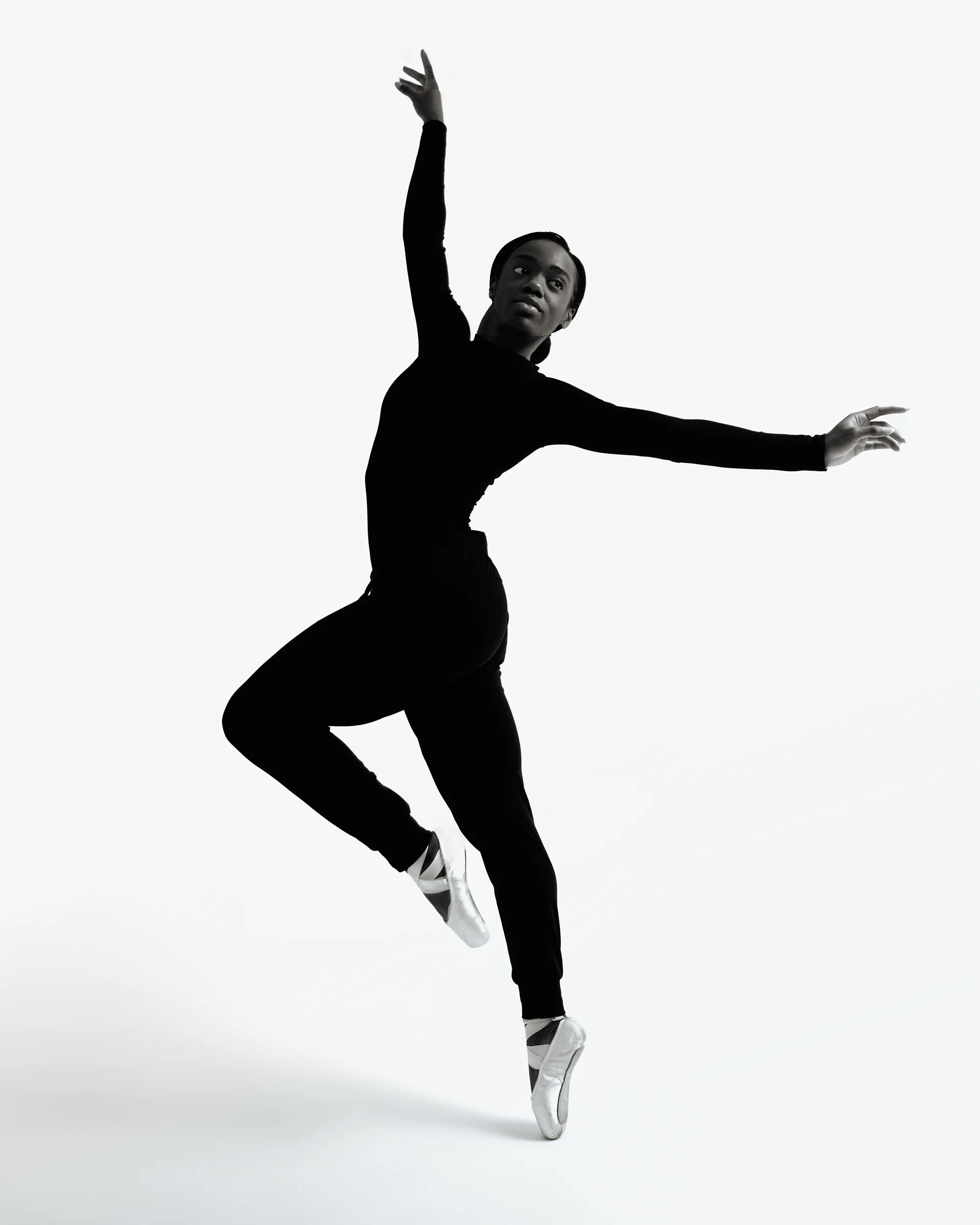 A dancer wearing all-black attire on pointe, captured mid-movement with arms gracefully extended against a white backdrop.