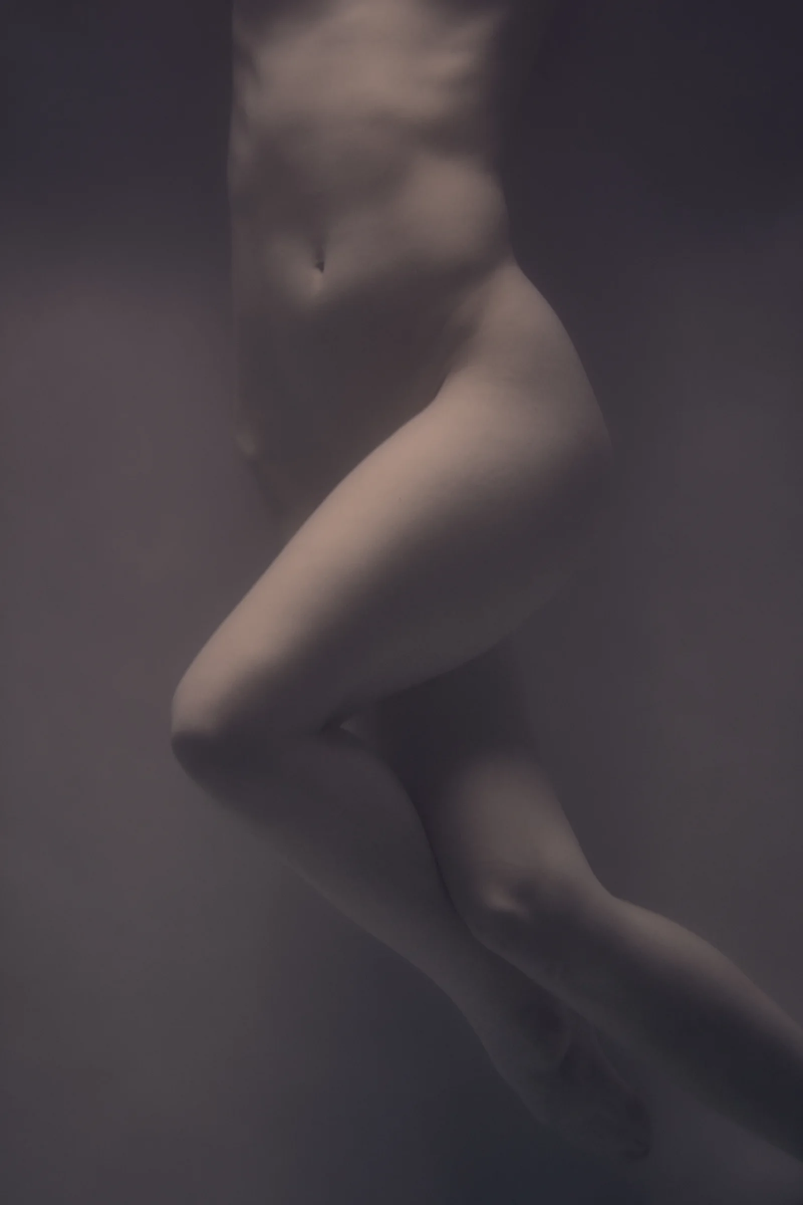 Softly lit, partially nude figure focusing on the torso and legs, captured in a minimal, subdued setting.