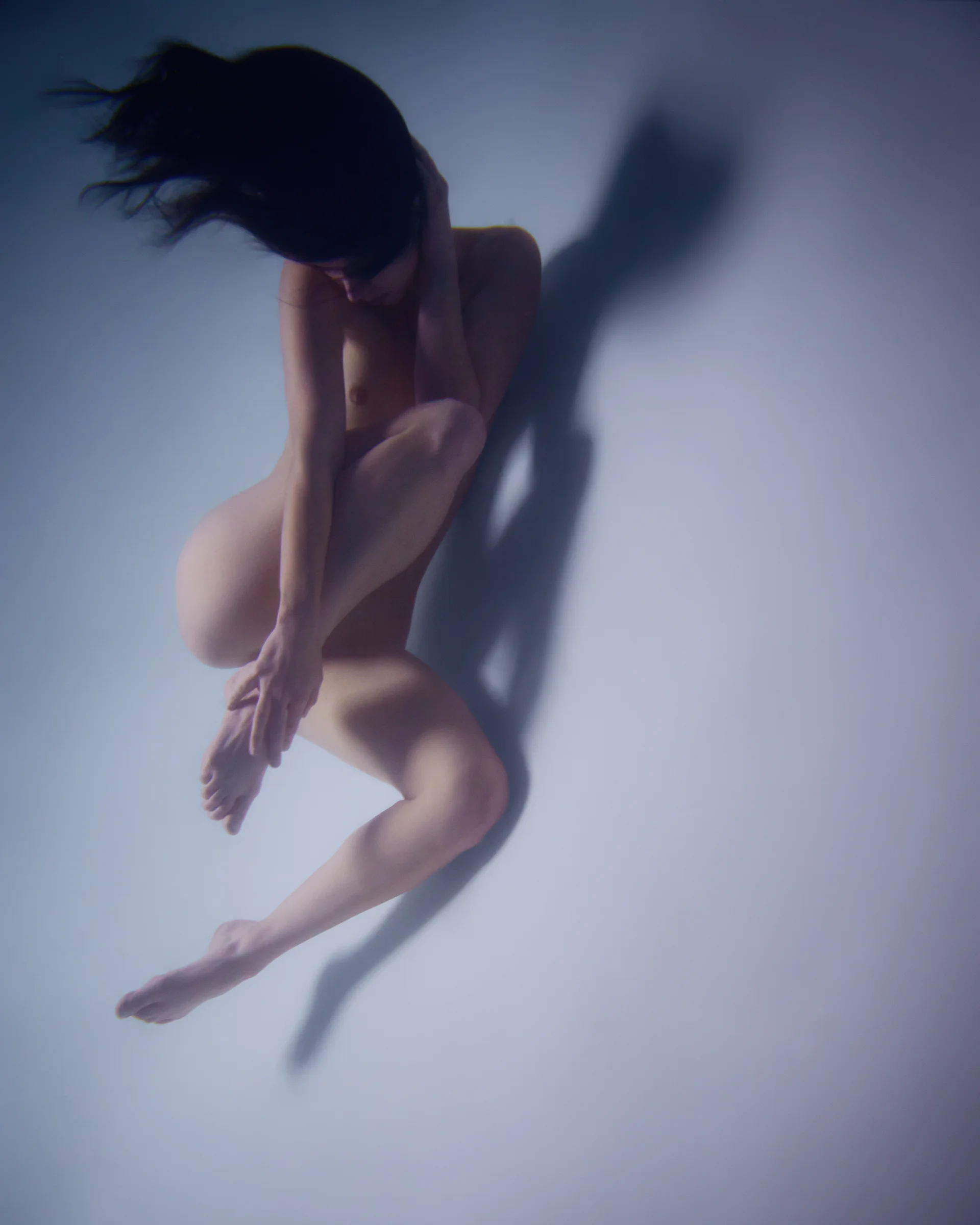 Partially nude figure posed as though floating, with hair flowing and a faint shadow cast on a pale background.