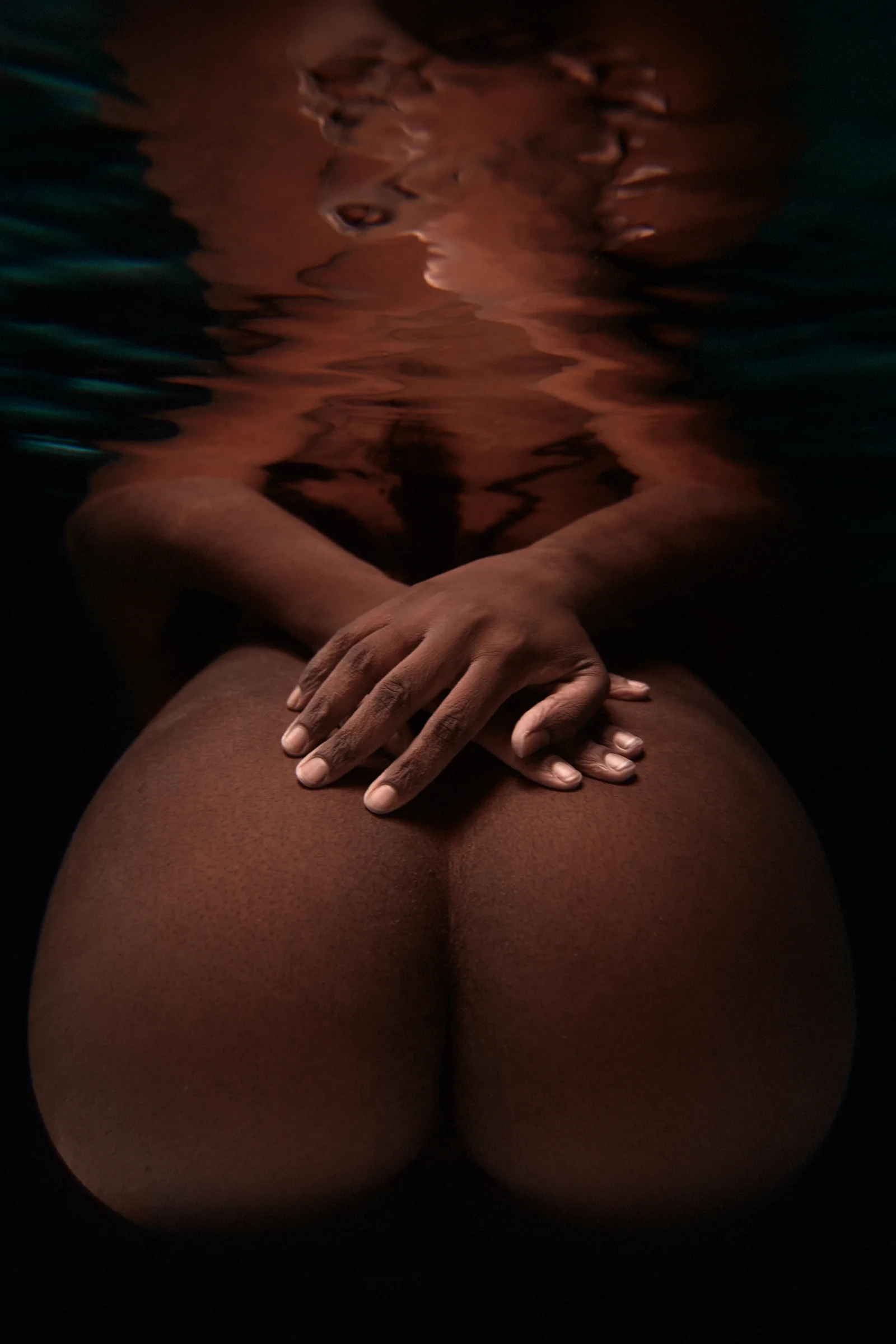 Underwater portrait of a partially nude individual with a dark complexion, hands folded over their knees and a distorted reflection on the water’s surface.