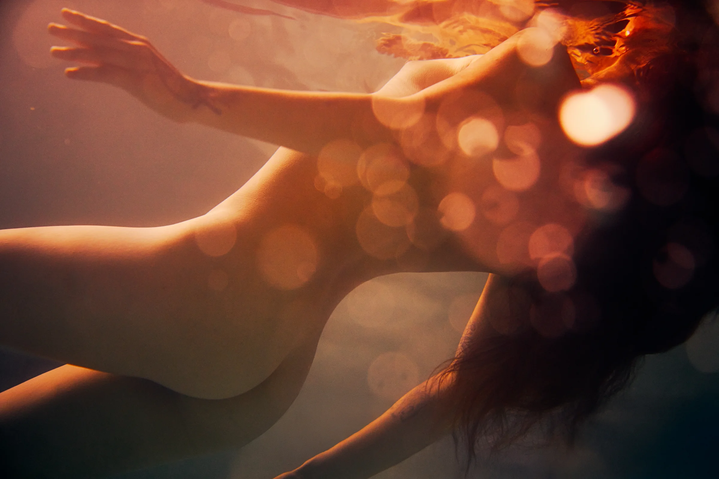 Underwater view of a partially nude figure with a warm, orange bokeh effect dancing across the scene.