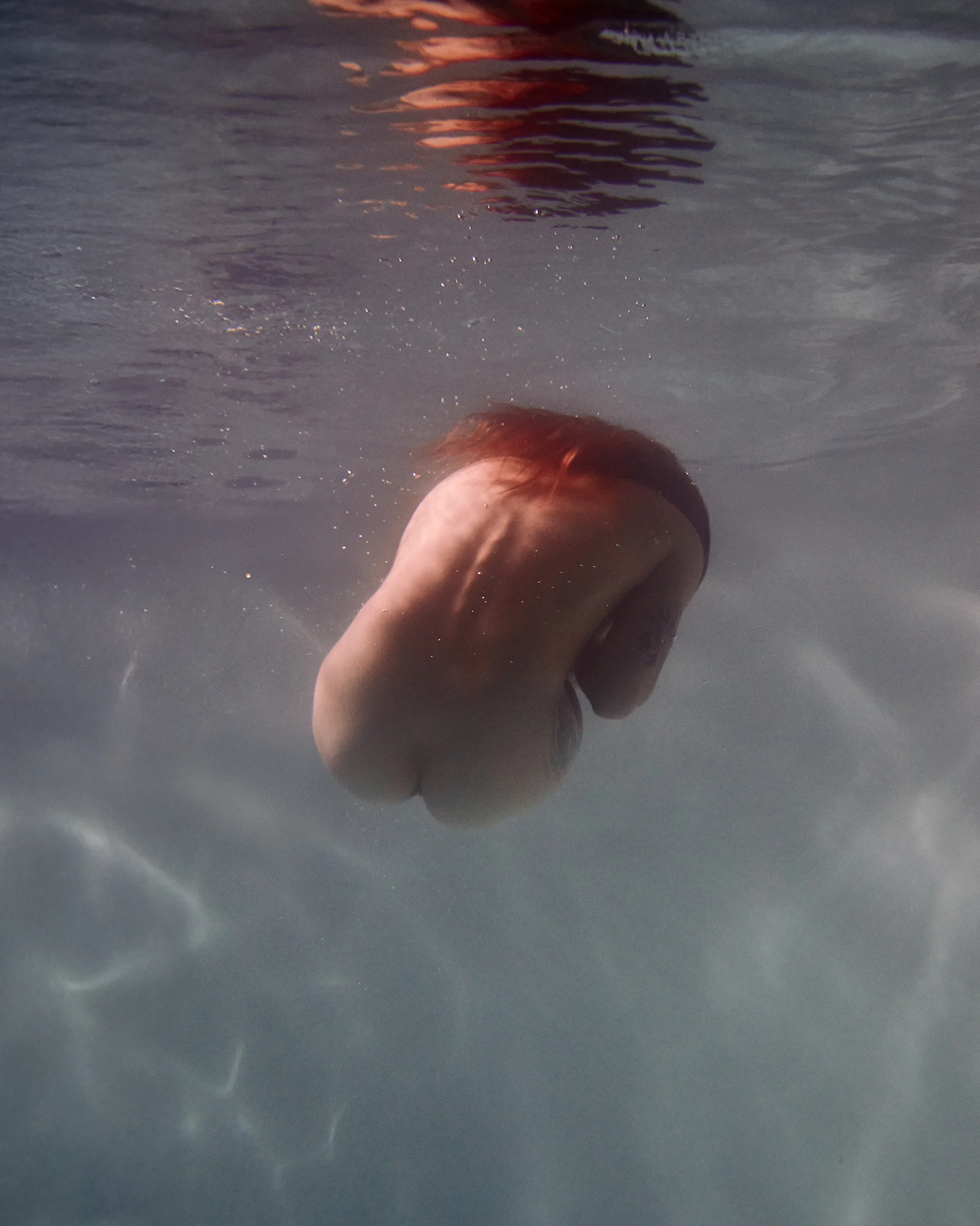 Underwater shot of a partially nude individual with red hair, captured mid-dive, as soft light shimmers around their back.